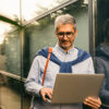 Homme portant des lunettes utilisant un ordinateur portable devant un bâtiment moderne en verre