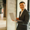 Un homme souriant en costume, tenant un ordinateur portable dans le couloir d'un bureau moderne.